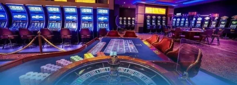 Bàn roulette và vòng quay trong sòng bài thật, chồng chip cạnh vòng quay, phía sau là dãy máy slot màn hình xanh